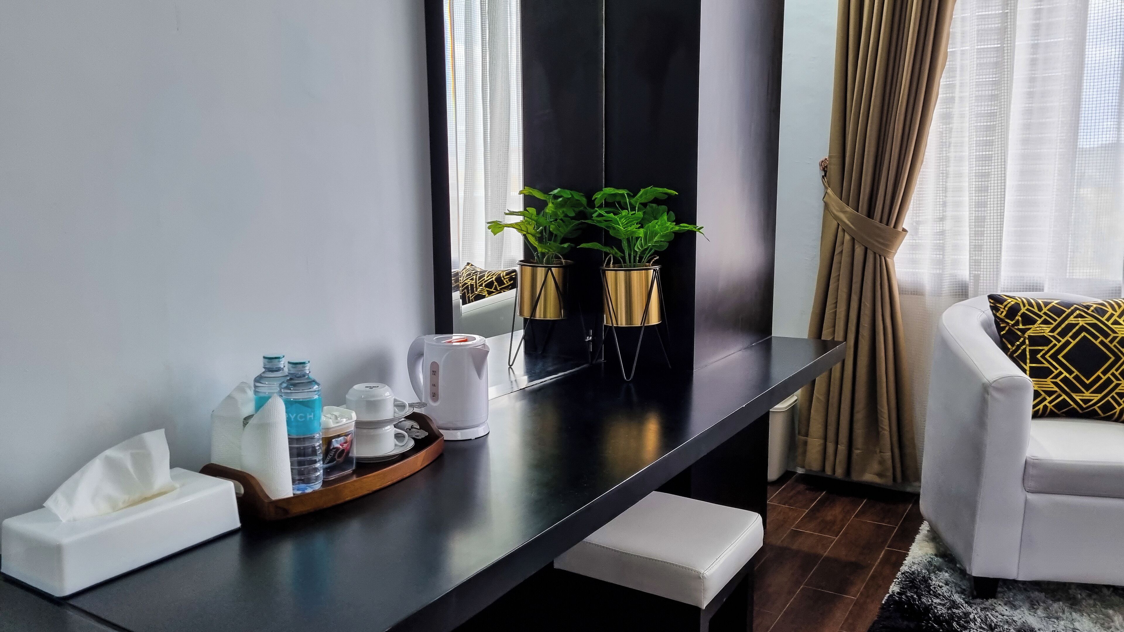 Et moderne hotelværelse med et mørkt træskrivebord, en hvid stol og en sofa med mønstrede puder. Der er to potteplanter på skrivebordet, en tissueæske, en vandflaske og en kaffemaskine.