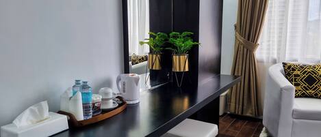 Una habitación de hotel moderna con un escritorio de madera oscura, una silla blanca y un sofá con cojines estampados. En el escritorio hay dos plantas en macetas, un caja de pañuelos, una botella de agua y una cafetera.