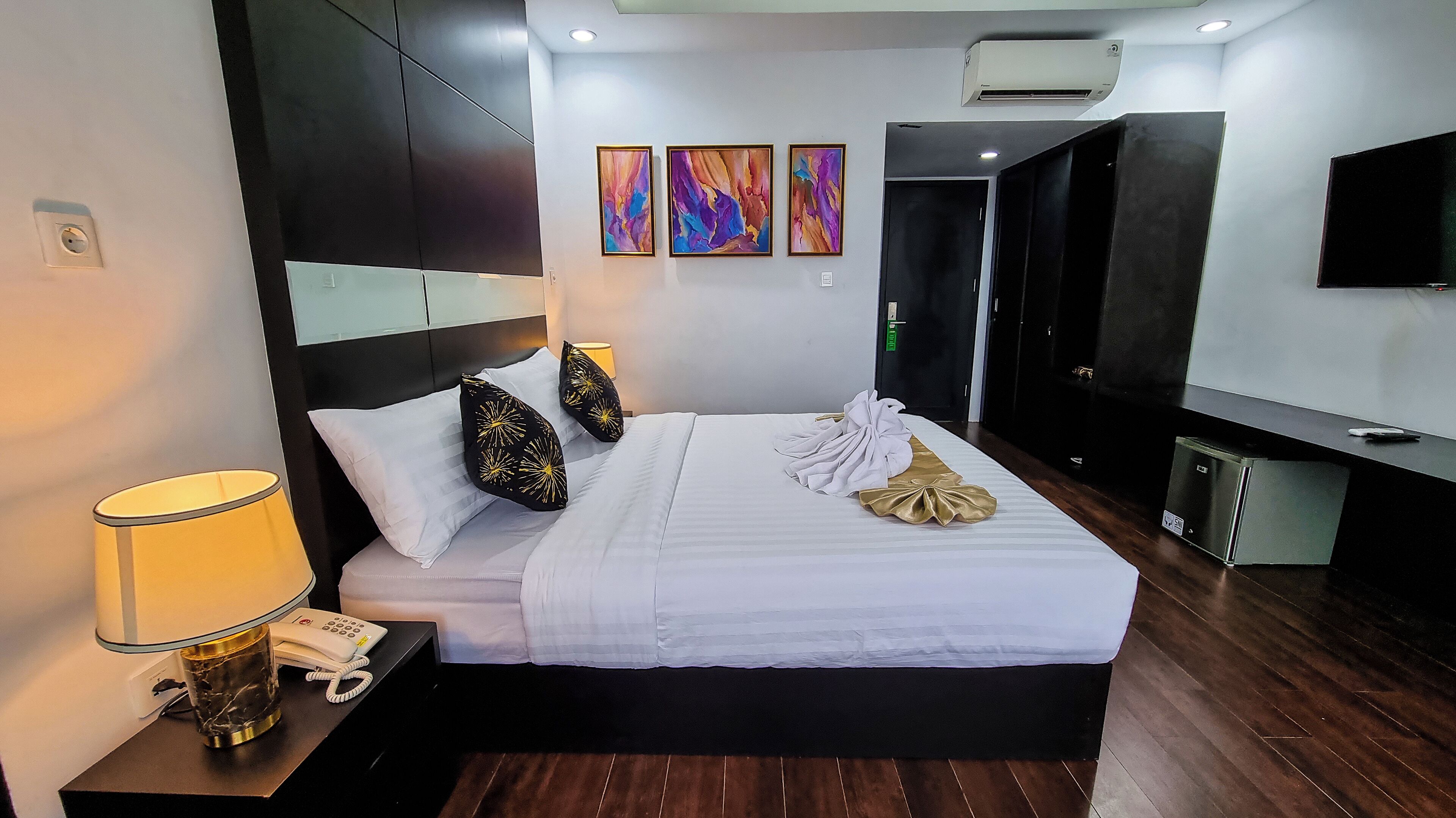 A hotel room with a bed, bedside lamp, phone, artwork on the wall, and a TV.