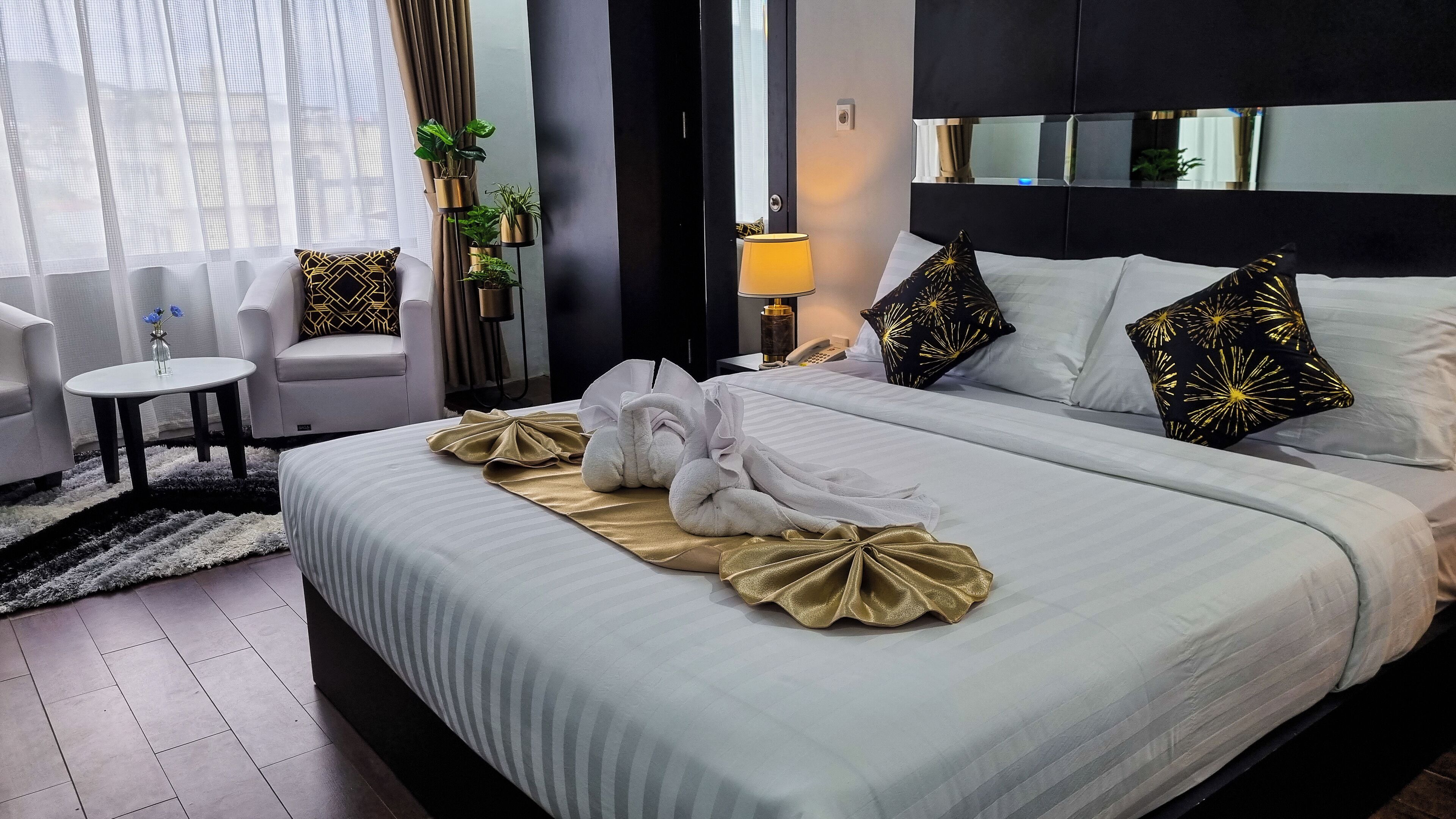 A neatly made bed with white linens and gold accents, a bedside table with a lamp, and a chair in a modern hotel room.