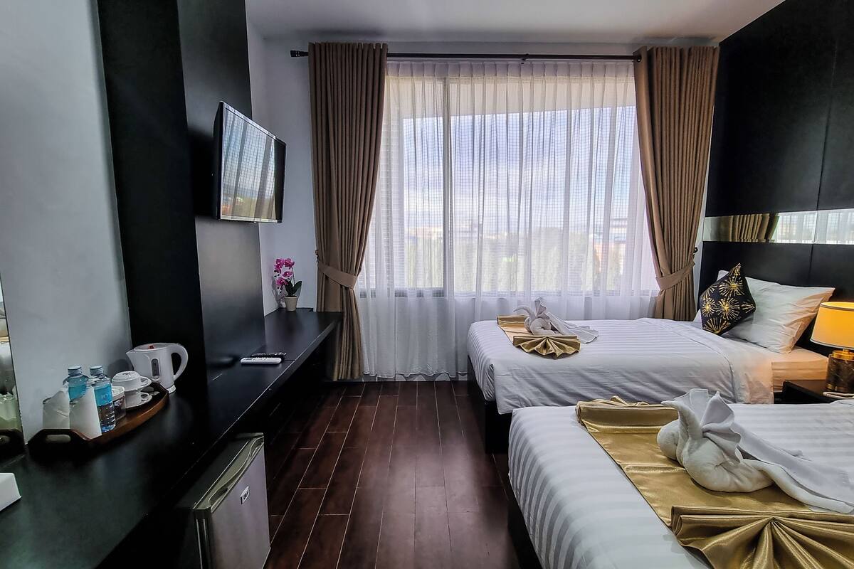 A hotel room with two beds, a TV, a desk with a kettle, and a window with curtains.