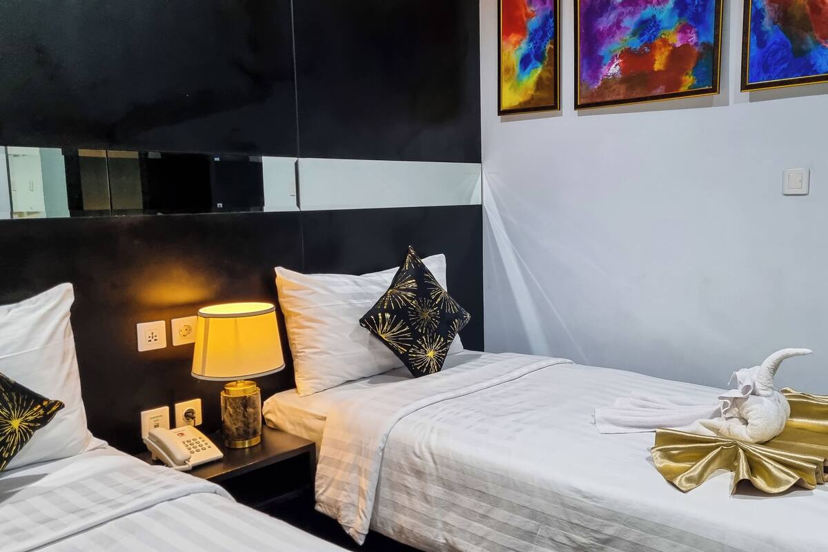 A hotel room with two beds, a bedside table with a lamp, a phone, and a decorative towel on the right bed.