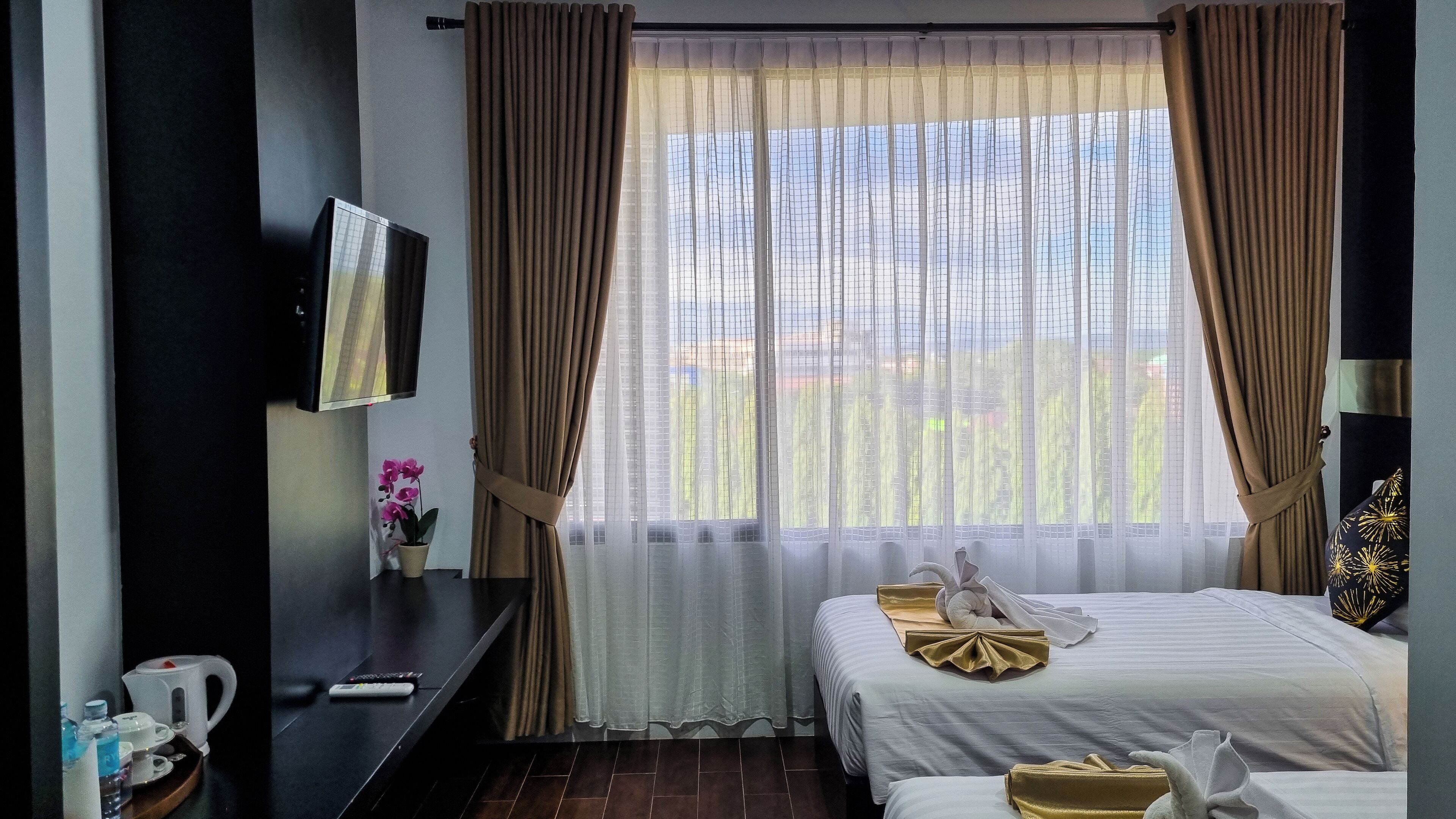 Una habitación de hotel con una cama, televisión, una mesita con una planta y una ventana con cortinas.