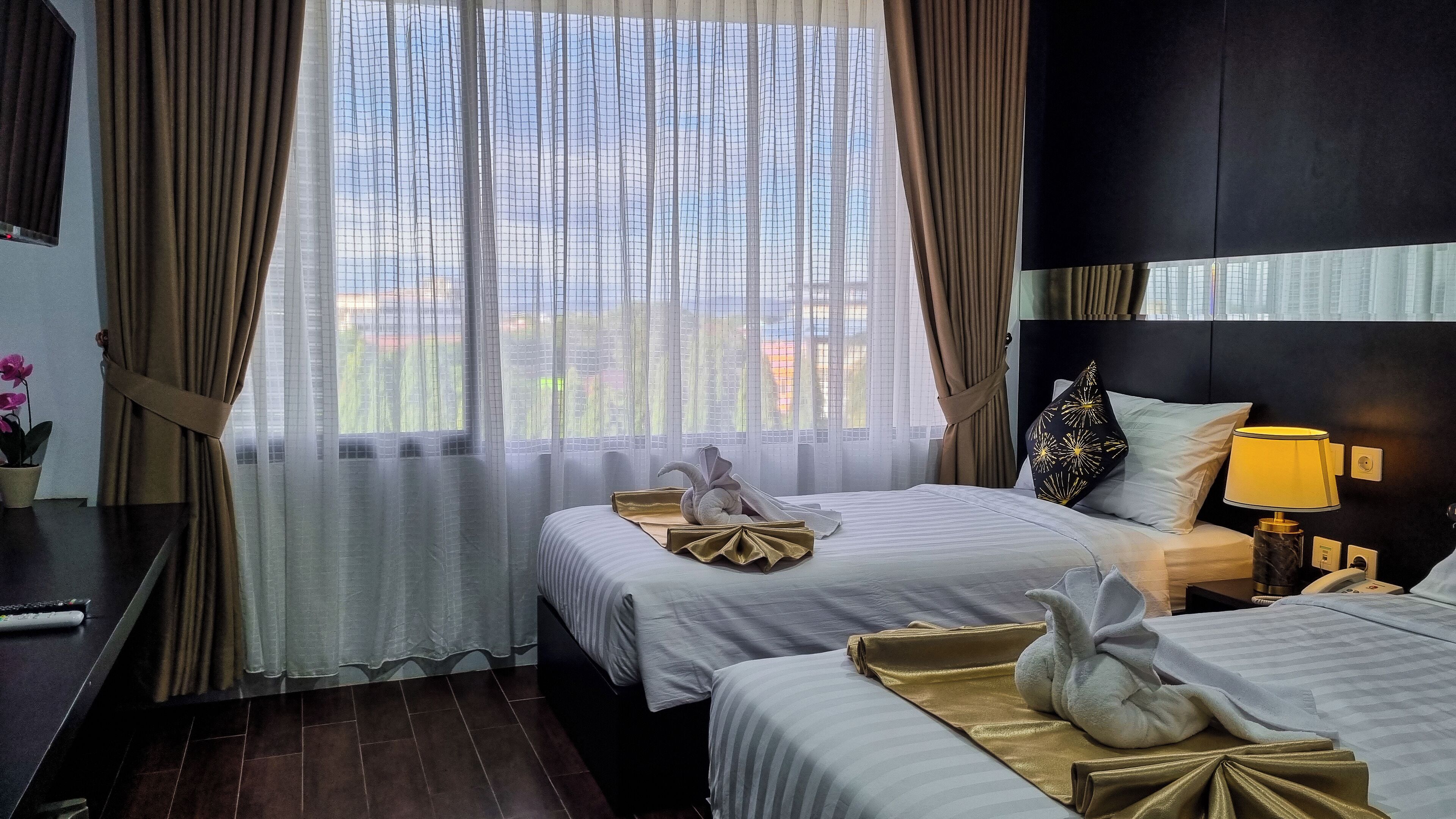 A hotel room with two beds, a nightstand with a lamp, and a view of greenery through sheer curtains.