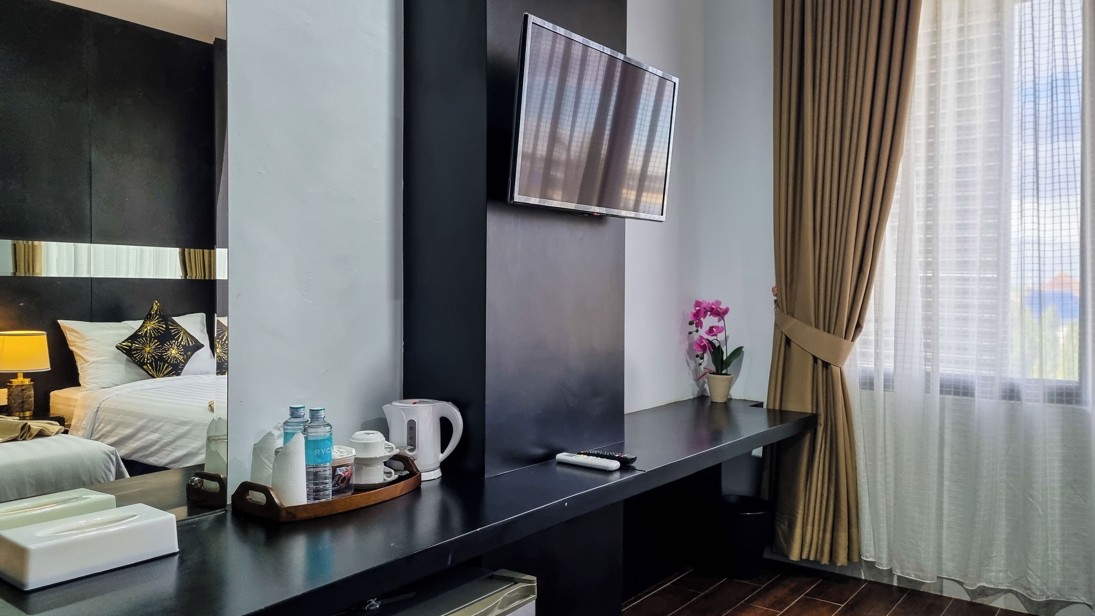 Quarto de hotel com cama, televisão fixada na parede, secretária com bandeja de produtos de higiene, orquídea em vaso e janela com cortinas.
