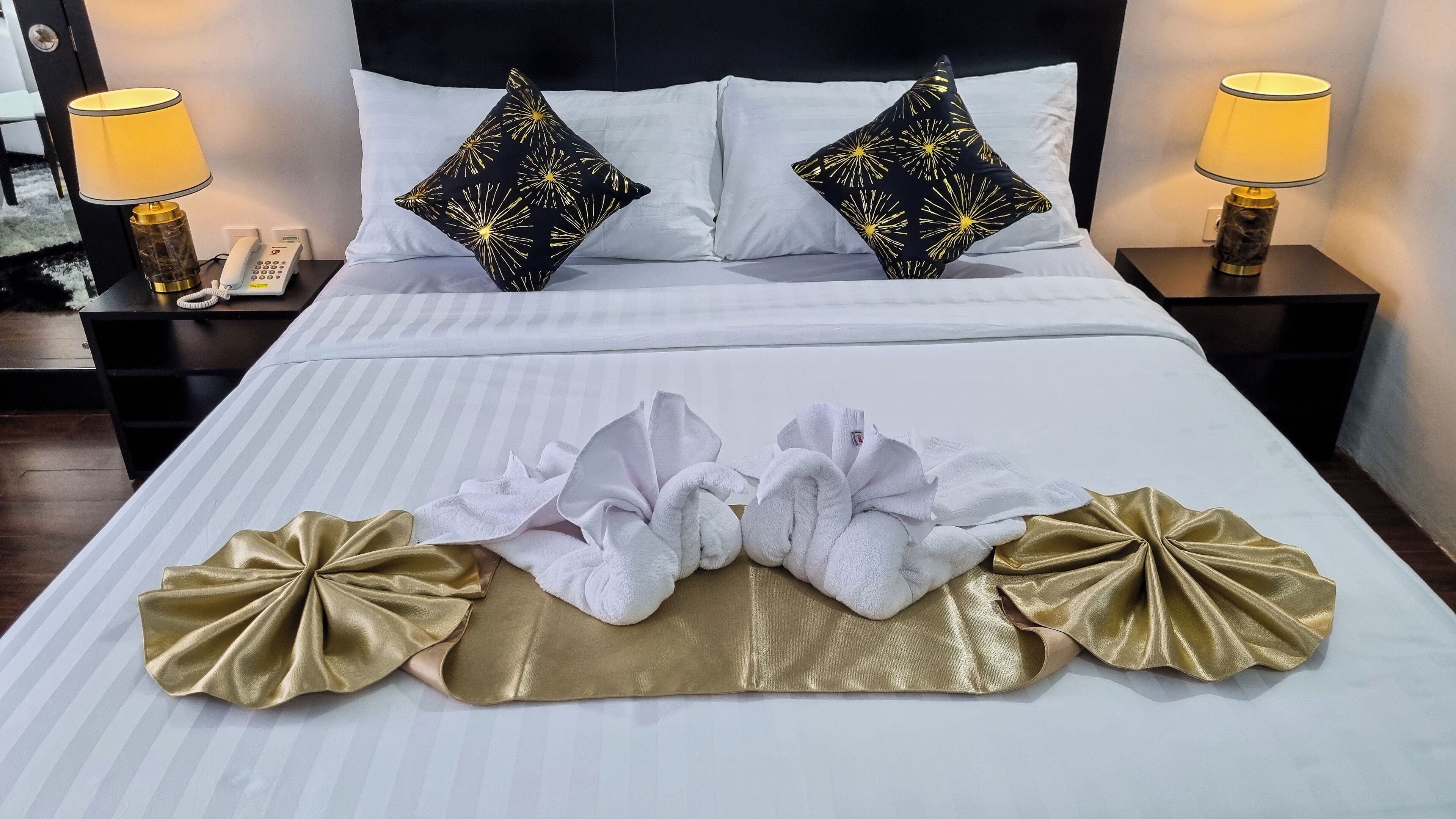 Una cama bien hecha con sábanas blancas, dos almohadas negras con un diseño de estrellas doradas y dos toallas con detalles dorados.