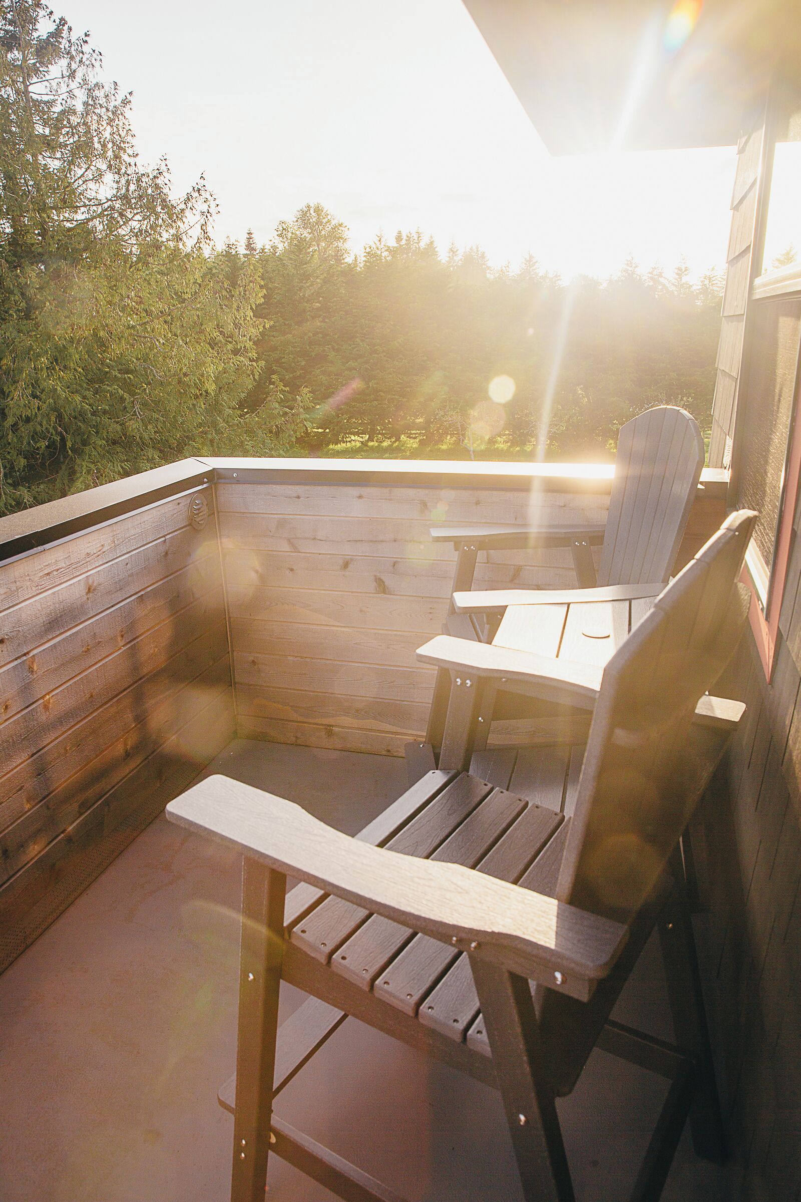 A wooden deck with a table and two Adirondack chairs, overlooking a forested area.
