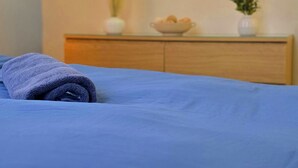 Une chambre avec une couvre-lit bleue, une commode en bois surmontée d’un tableau, et une petite plante posée sur la commode.