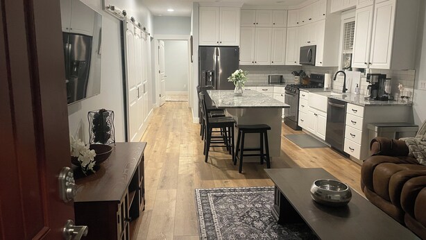 Una cocina moderna con armarios blancos, electrodomésticos de acero inoxidable, una isla central con taburetes y una mesa de comedor de madera con un tazón.