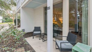 A covered outdoor seating area with two chairs and a table, a building number 230, and a view of a lit interior through a glass door. - Salt Air Nook - Pool Access (Jekyll Island)