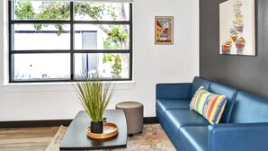A modern living room with a blue sofa, a potted plant on a black table, and a window with blinds. - King Suite & Twin Room | Condo at The Well Mouratoglou | Tennis & Pickleball (Zephyrhills)