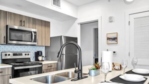 A modern kitchen with stainless steel appliances, a double sink, and a dining area set for four. - King Suite & Twin Room | Condo at The Well Mouratoglou | Tennis & Pickleball (Zephyrhills)