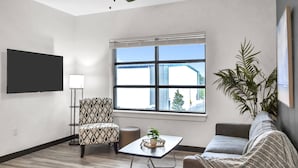 A modern living room with a flat-screen TV, a patterned armchair, a small table with a plant, a grey sofa, and a large window with blinds.