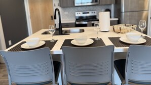 A modern kitchen with a dining table set for four, featuring a central island with a bowl and wine glasses.
