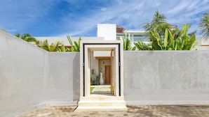 A modern building entrance with a concrete facade, a brick pathway leading to a wooden door, and tropical plants in the background. - Villa Casa Siesta by Pertama Stay (Ungasan)