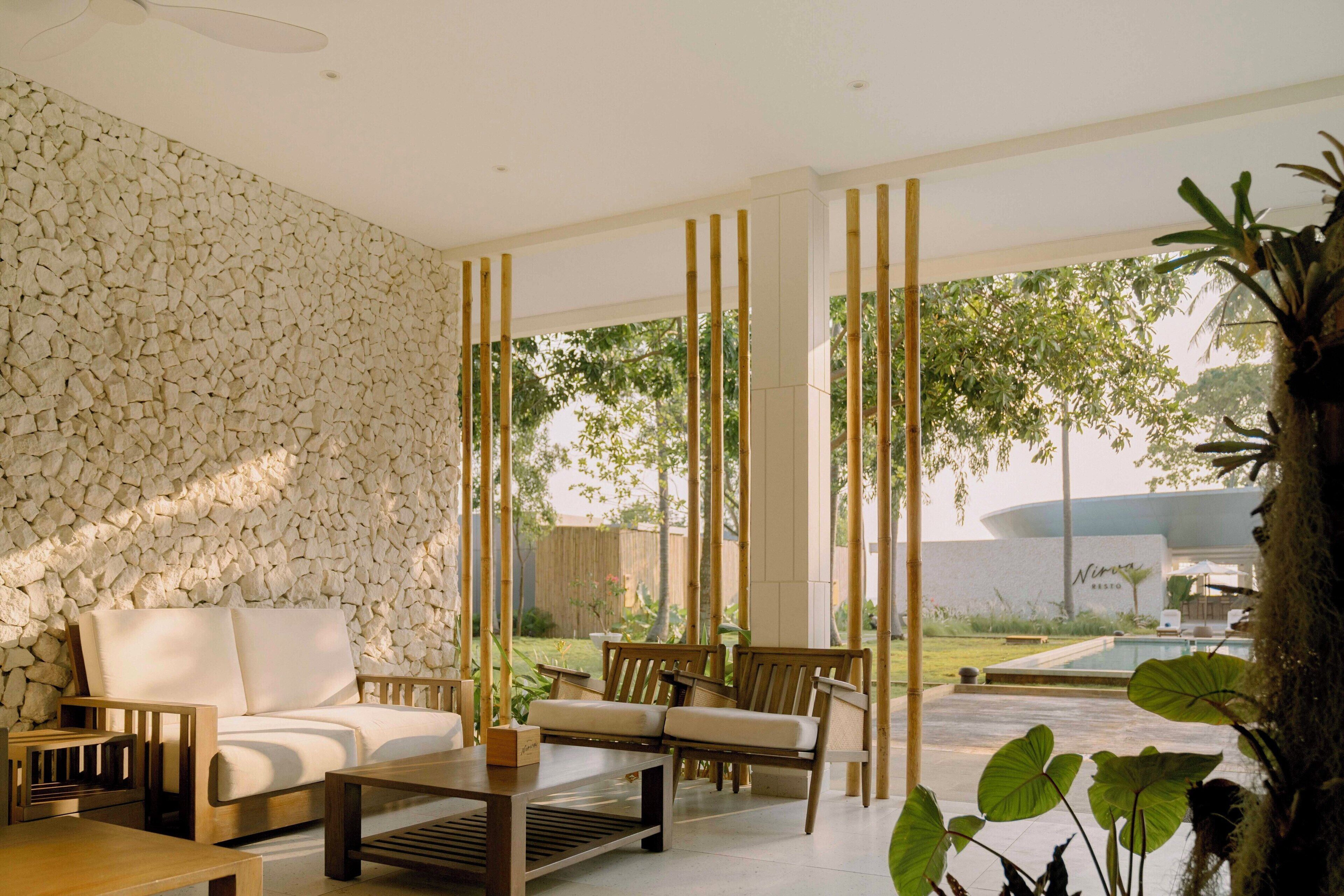 A modern outdoor seating area with a stone wall, wooden furniture, and a view of a pool and greenery.