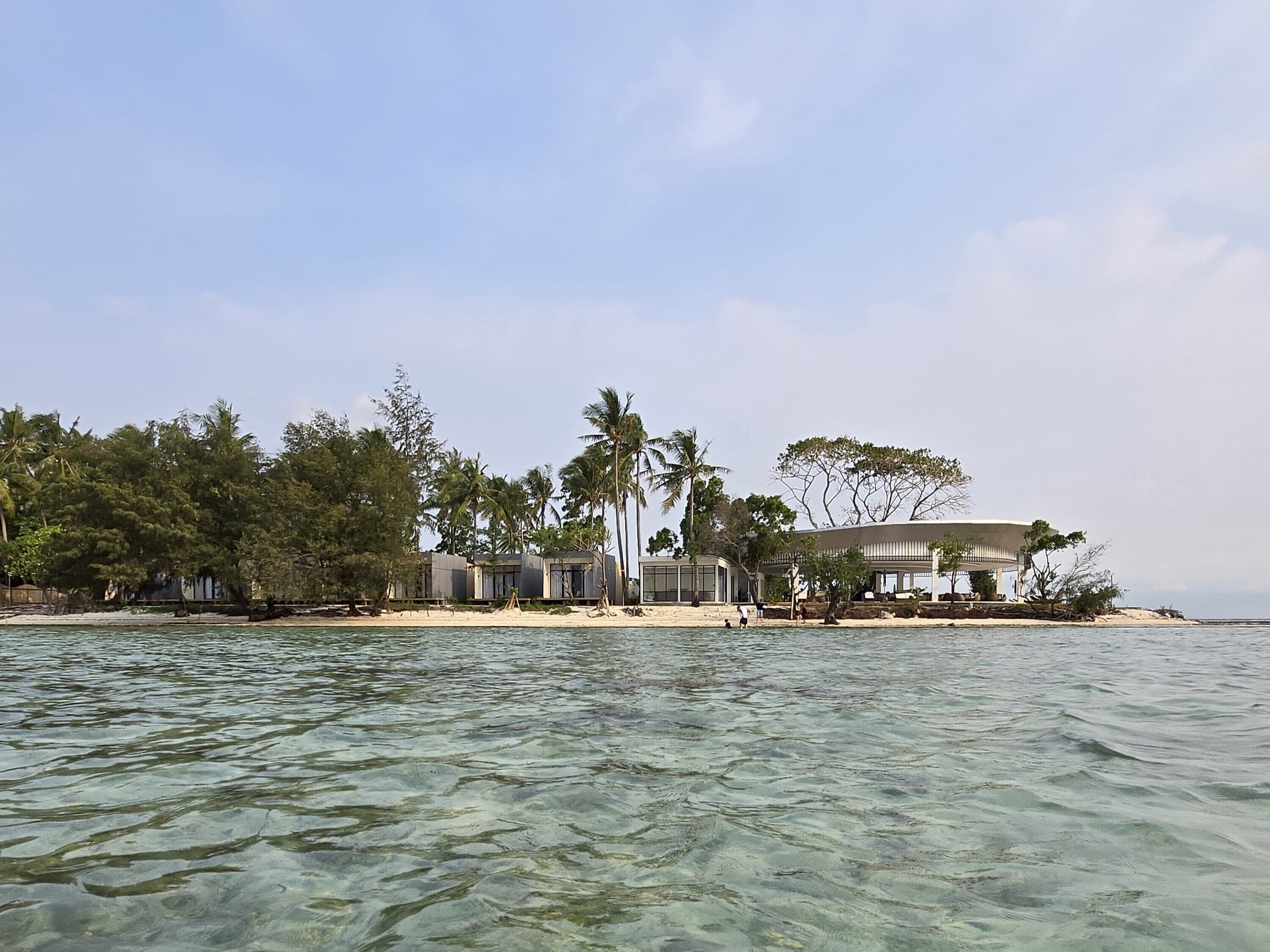 A beachfront property with multiple buildings, clear water, and palm trees.