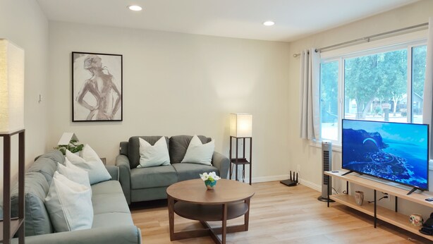 A living room with a grey sofa, a round wooden coffee table, a flat-screen TV, and a window with a view of trees. - 261-6 Cozy 2-Bedroom Apartment near Downtown Mountain View (Mountain View)