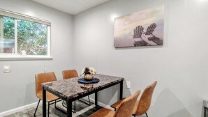 Uma área de refeições com uma mesa de mármore, quatro cadeiras marrons, um vaso com flores, um quadro com cadeiras e uma janela com vista para árvores.
