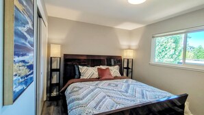 Quarto com cama, duas luminárias de cabeceira, janela com vista para árvores e um quadro na parede.