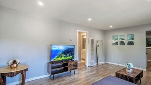 Uma sala de estar com televisão de tela plana, piso de madeira, uma mesa redonda pequena com um vaso e um sofá.