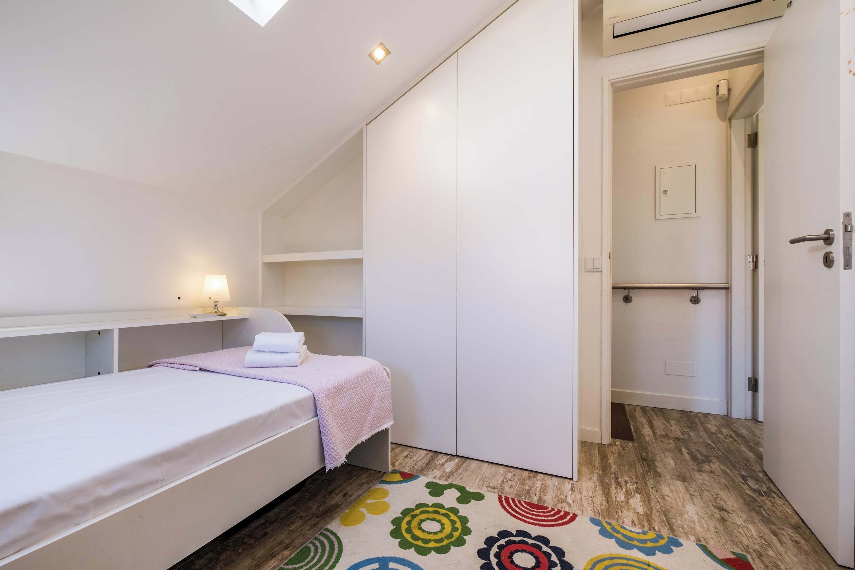 A small bedroom with a single bed, a bedside table with a lamp, a wardrobe, and a door leading to a bathroom.