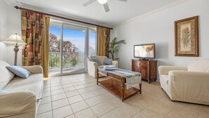 Una sala de estar con sofá, sillón, mesa de centro y televisión. Hay un balcón con cortinas y ventilador de techo.