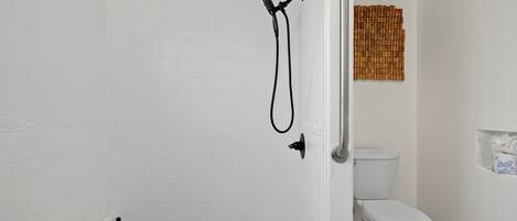 Un baño con paredes de azulejos blancos, piso de baldosas color beige, inodoro blanco, ducha con cabezal de mano y un perchero con una toalla.