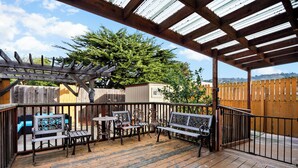 Una terraza techada al aire libre con piso de madera, barandales de metal y mobiliario exterior que incluye bancos y mesas.