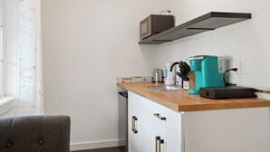 Cocina compacta con armarios blancos, encimera de madera, microondas, cafetera y ventana con cortina blanca.