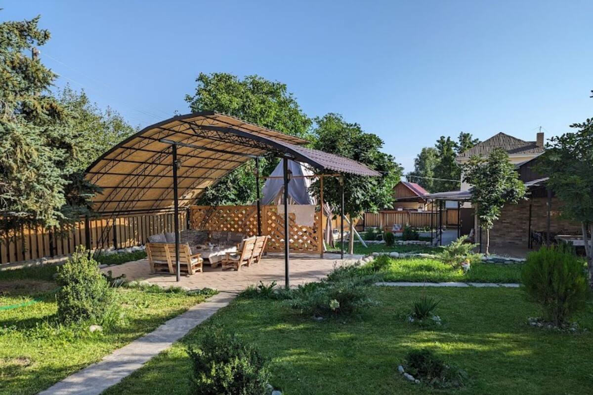 Un espace extérieur aménagé, partiellement abrité, avec un foyer, entouré d’une clôture en bois et de verdure.