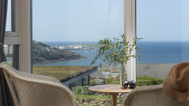 Un balcon agrémenté de chaises en osier, une petite table ronde avec deux verres à vin et un vase rempli de verdure, offrant une vue sur la côte.