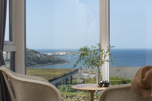 A balcony with wicker chairs, a small round table with two wine glasses and a vase with greenery, overlooking a coastal view.