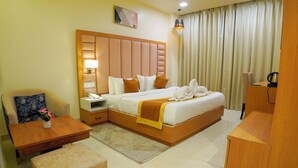 A hotel room with a large bed, a bedside table, a chair, a small table with a vase, and a wooden desk. - Hotel Cheer Royale (Bareilly)