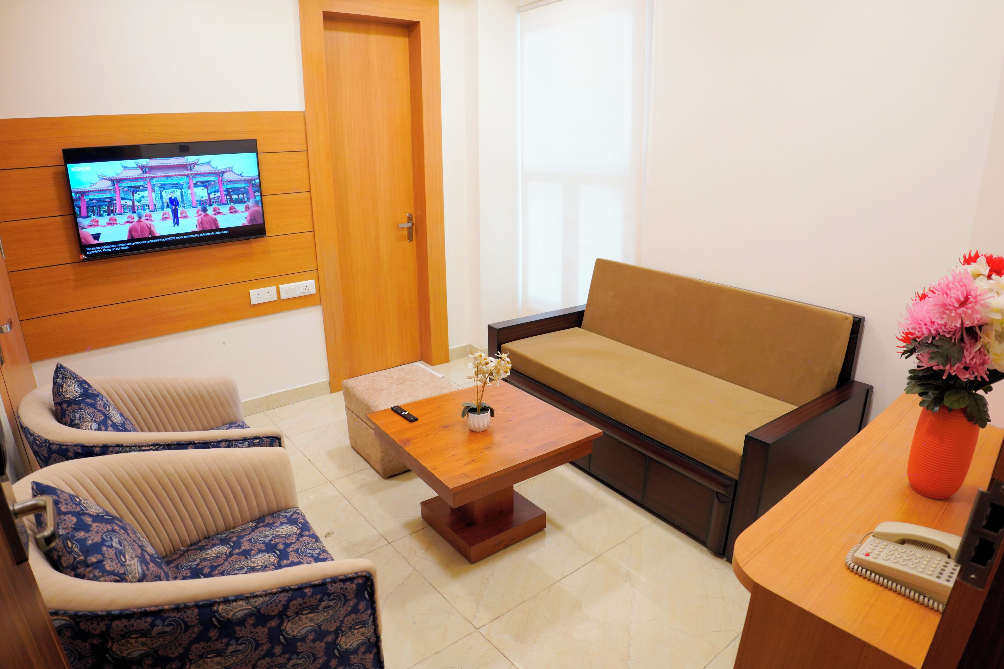 A living room with a sofa, two armchairs, a coffee table, a TV mounted on the wall, and a vase with flowers on a side table.