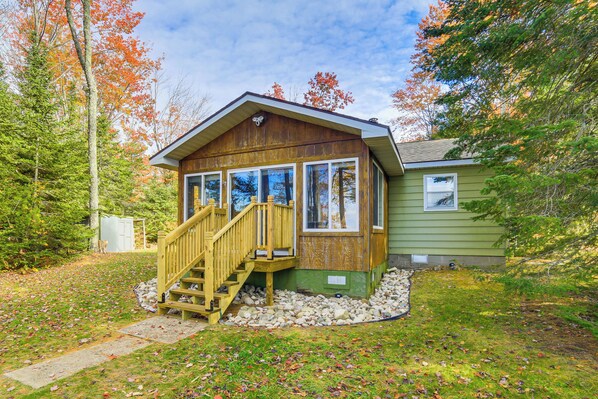 A cabin with a wooden staircase leading to a deck, surrounded by trees and a stone pathway. - Dock & Fire Pit: Lakefront Cottage in Gould City! (Gould City)