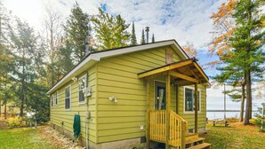 A small, single-story cabin with a porch, situated next to a body of water, surrounded by trees. - Dock & Fire Pit: Lakefront Cottage in Gould City! (Gould City)