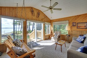 A spacious room with a large window overlooking a body of water, a ceiling fan, and a cozy seating area with a sofa and chairs. - Dock & Fire Pit: Lakefront Cottage in Gould City! (Gould City)