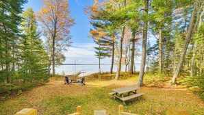 A wooden deck leading to a picnic area with benches and a table, surrounded by trees and a lake. - Dock & Fire Pit: Lakefront Cottage in Gould City! (Gould City)