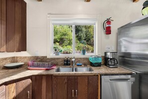 A kitchen with a stainless steel dishwasher, a coffee maker, and a window with a view of greenery. - Hot Tub Retreat Near Russian River • BBQ • Family Friendly (Guerneville)