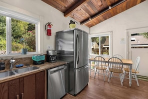 A modern kitchen with a stainless steel refrigerator, a coffee maker, and a dishwasher. There is a window with a view of greenery outside. - Hot Tub Retreat Near Russian River • BBQ • Family Friendly (Guerneville)