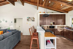 A spacious kitchen with wooden cabinets, a central island, and a dining area with a grey sofa and metal bar stools. - Hot Tub Retreat Near Russian River • BBQ • Family Friendly (Guerneville)