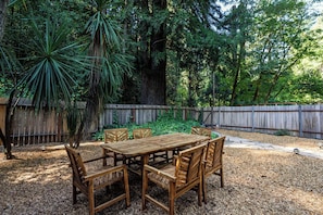 Un patio al aire libre con una mesa y sillas de madera, rodeado por una valla de madera y abundante follaje.
