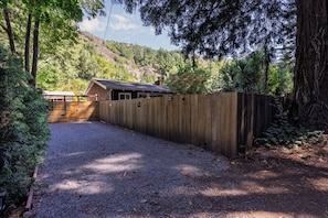 A gravel driveway leading to a wooden cabin with a metal roof, surrounded by dense forest and hills. - Hot Tub Retreat Near Russian River • BBQ • Family Friendly (Guerneville)