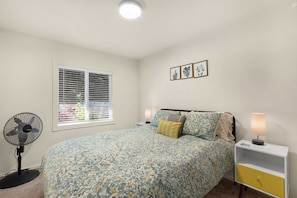 A bedroom with a bed, bedside lamps, a standing fan, a window with blinds, and framed botanical prints on the wall. - Hot Tub Retreat Near Russian River • BBQ • Family Friendly (Guerneville)