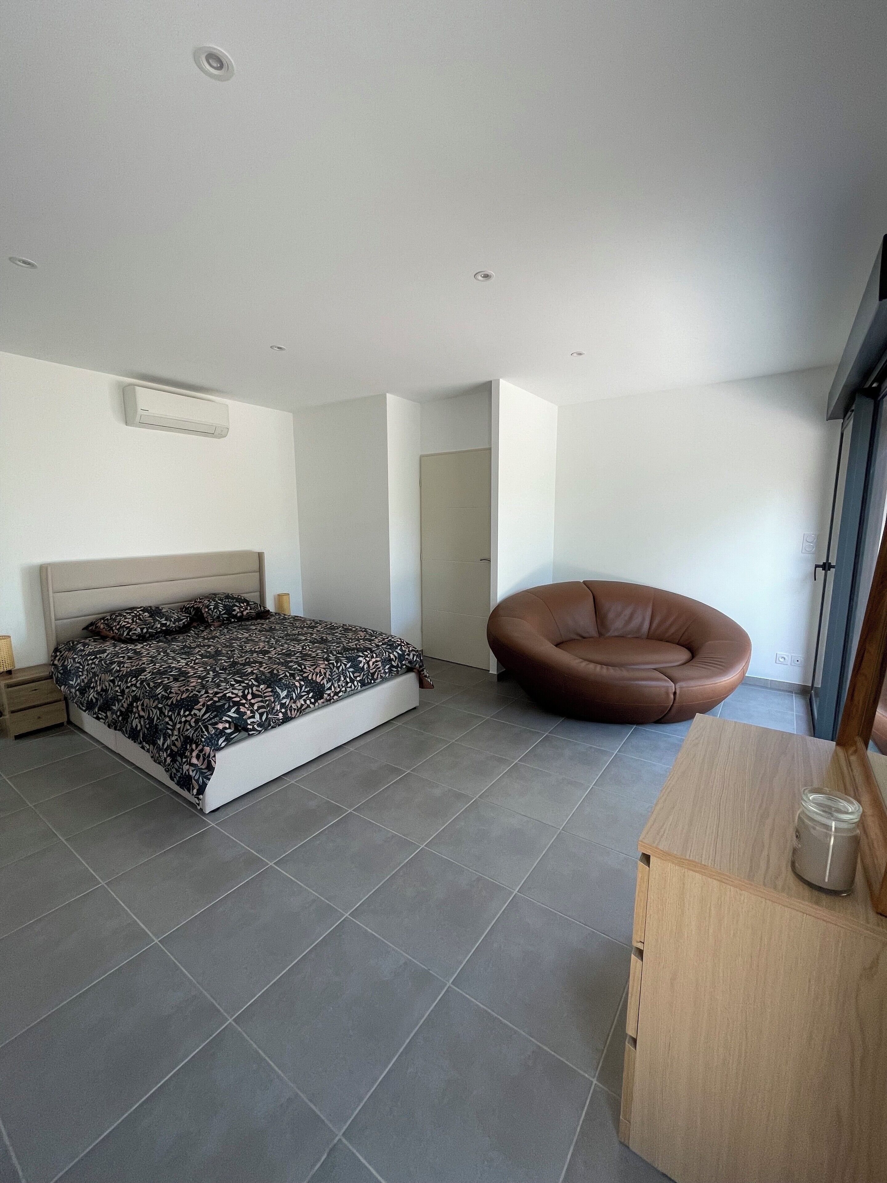 Ein modernes Schlafzimmer mit einem Bett, einem braunen Ledersessel, einem Nachttisch aus Holz und einem kleinen Holztisch mit einem Einmachglas.