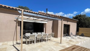 An outdoor patio with a wooden deck, a pergola with white fabric, and a dining area with chairs and tables.