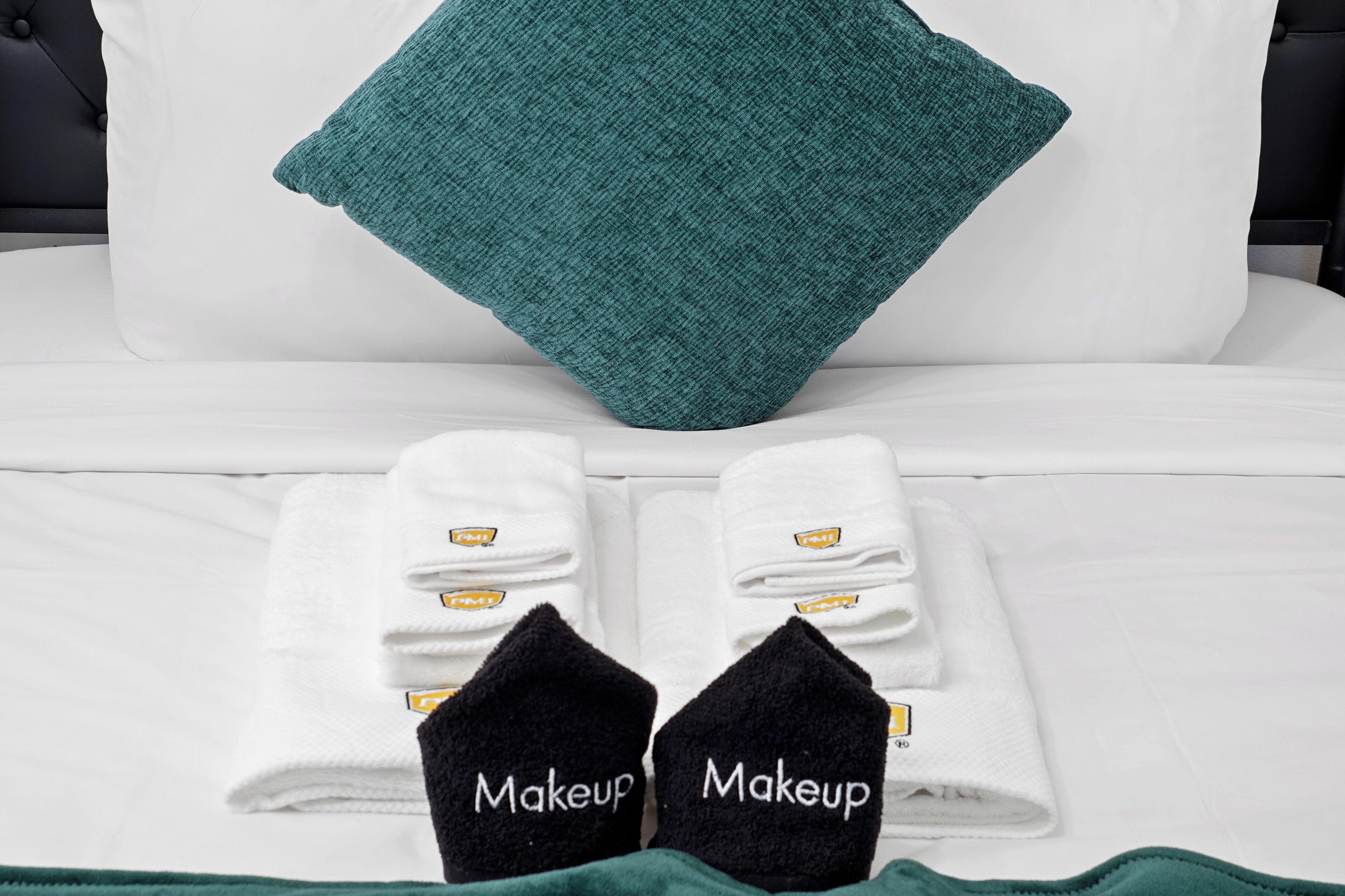 Una cama bien tendida con ropa de cama blanca y aguamarina, dos toallas con la palabra 'Makeup' escrita y un cojín decorativo aguamarina.
