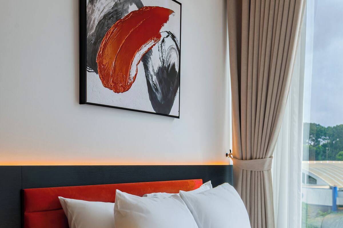 Ein Hotelzimmer mit einem modernen Bett, einem Kopfteil, einem gerahmten abstrakten Gemälde und Blick auf eine Brücke durch das Fenster.