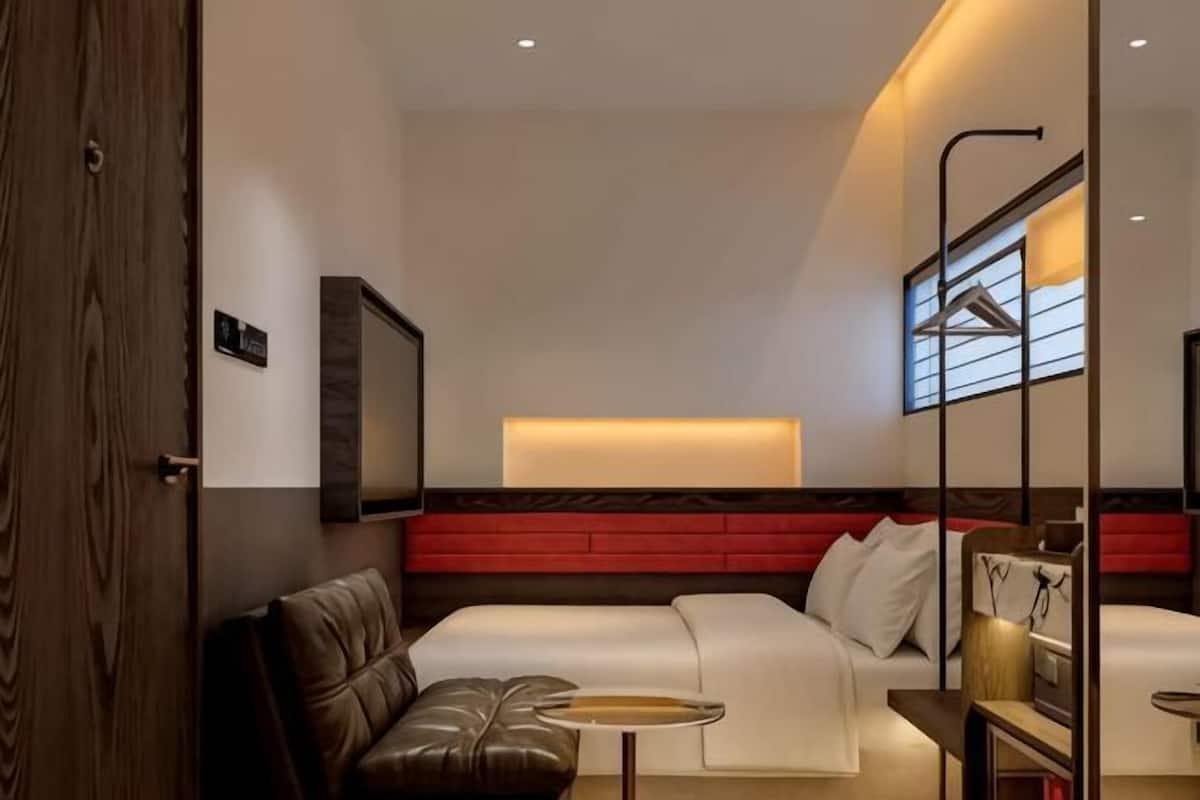 Ein modernes Hotelzimmer mit Bett, Sitzgelegenheit, Fernseher und einem Fenster mit Jalousien.